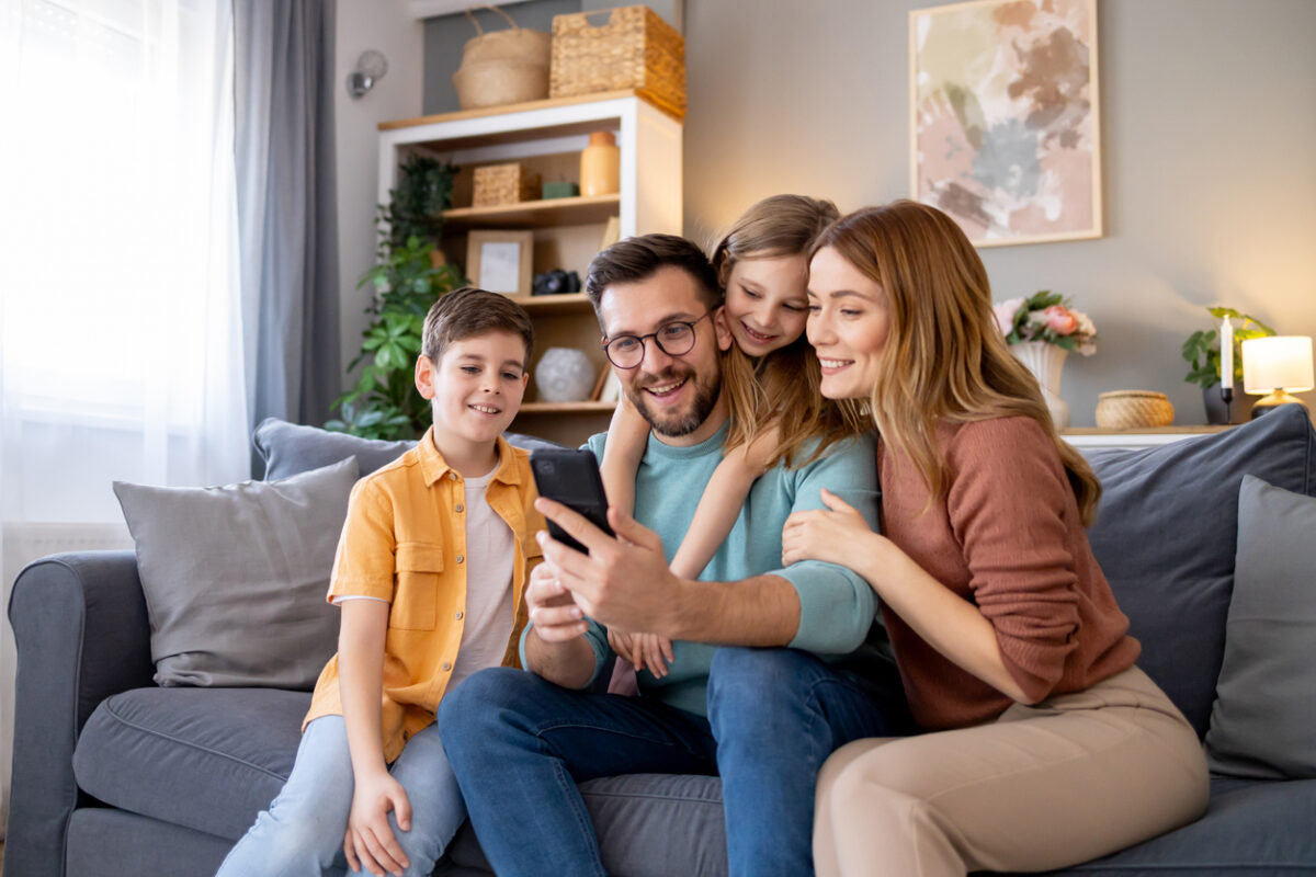 Capturing a cosy, home scene where a happy family with a mother, father, and kids engaging with a mobile phone, enjoying their time together