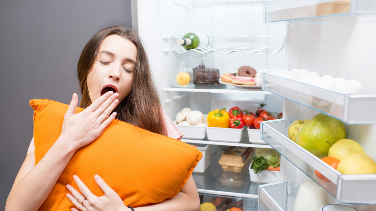 Young woman with pillow yawning standing in front of the refrigerator. Concept of eating during the night