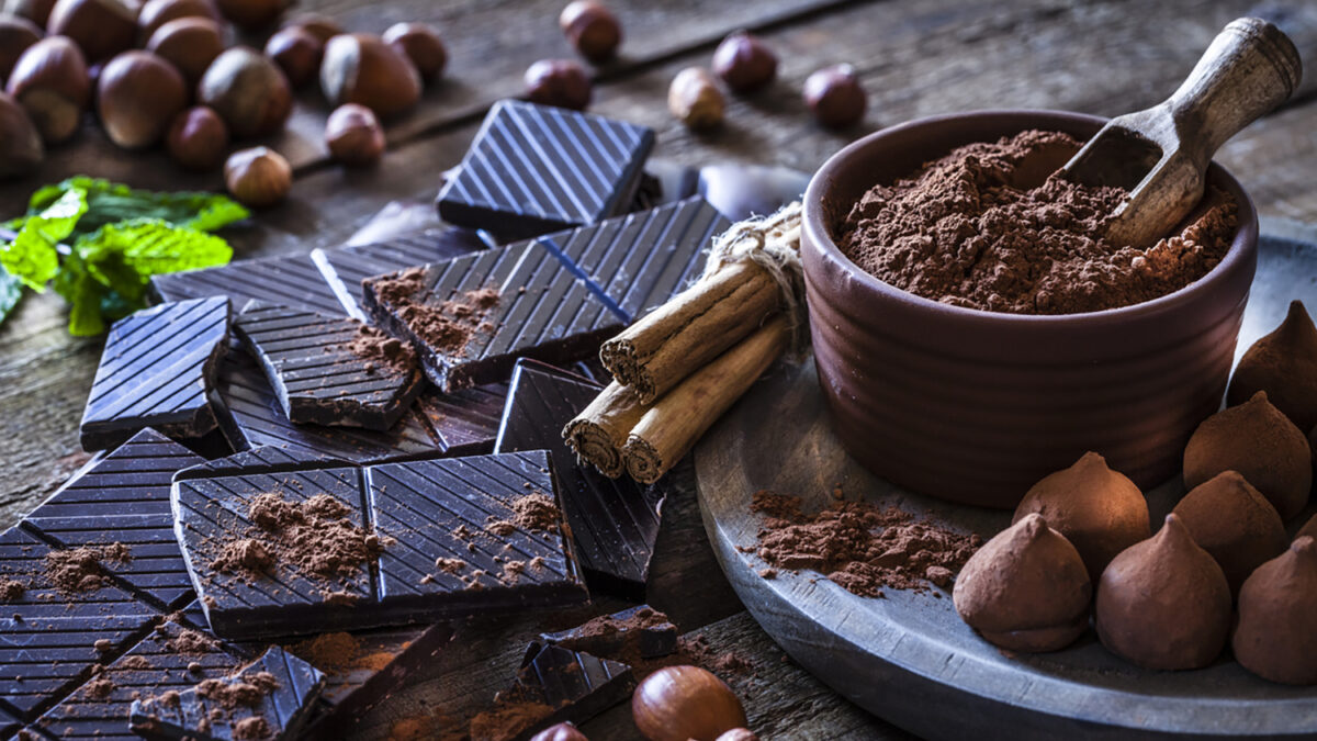 Rustic wooden table filled with ingredient for preparing homemade chocolate truffles. The composition includes dark chocolate truffles, chocolate bars, cocoa powder, cinnamon sticks, vanilla beans, mint leaves and hazelnuts. Predominant color is brown.