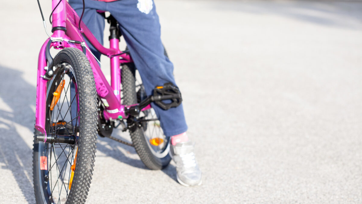 Anonymous young elementary school age child, girl sitting on a bike, legs closeup, front view, copy space background. Kid sitting on a bicycle, means of transport, healthy physical activity concept