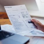 A person reviews their energy bill while using a laptop at home. The document includes various charges and electricity usage information.
