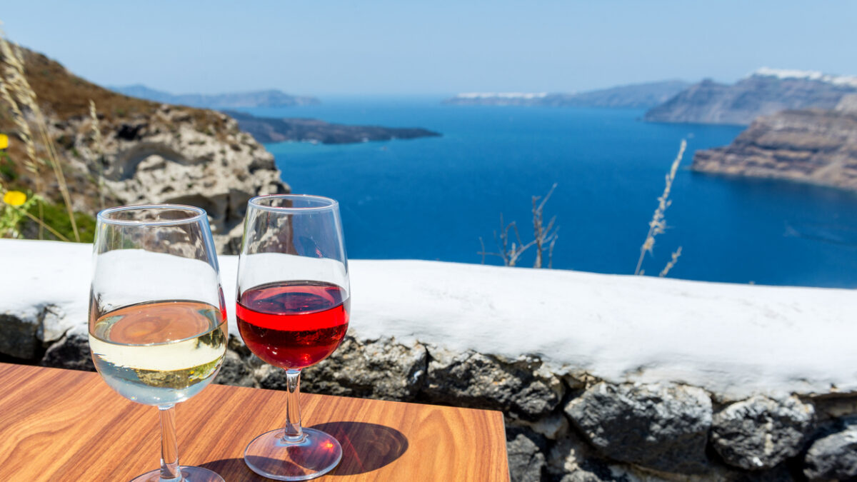 Horizontal color image of two glasses of wine - white wine & rose wine. Aegean sea and volcano in the background