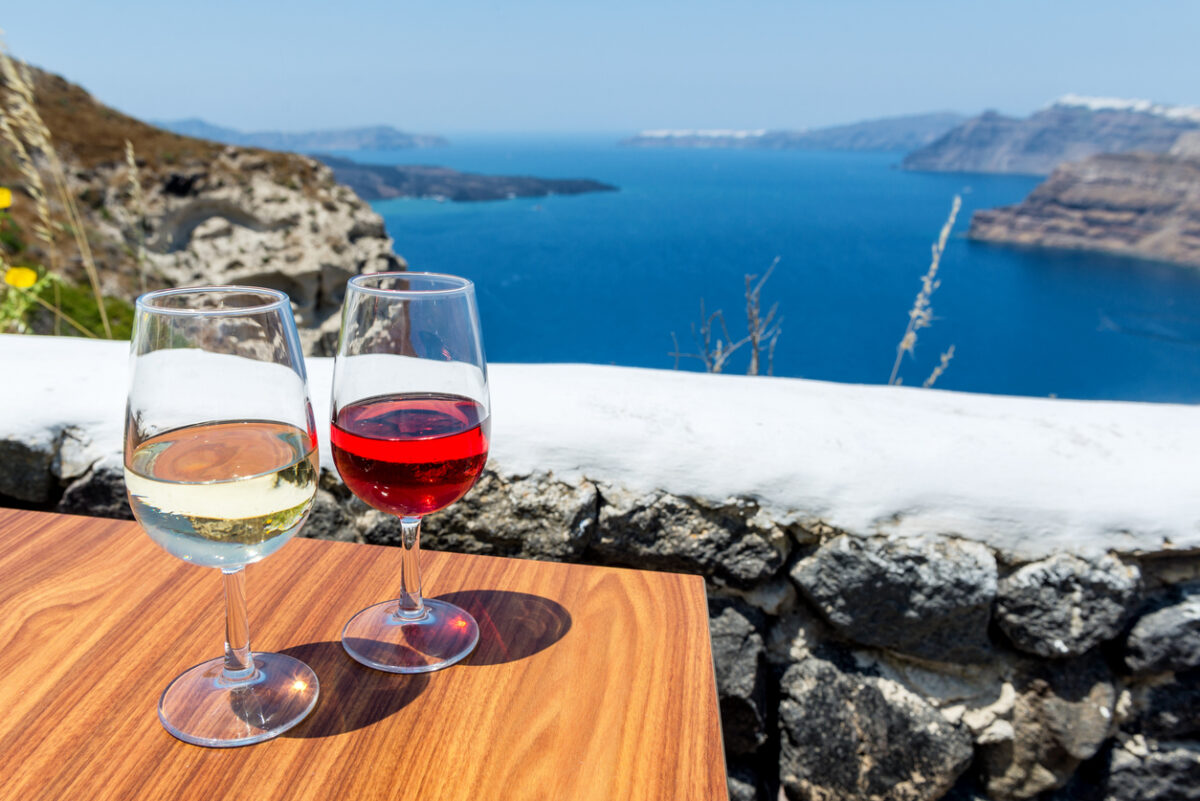 Horizontal color image of two glasses of wine - white wine & rose wine. Aegean sea and volcano in the background
