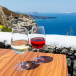 Horizontal color image of two glasses of wine - white wine & rose wine. Aegean sea and volcano in the background