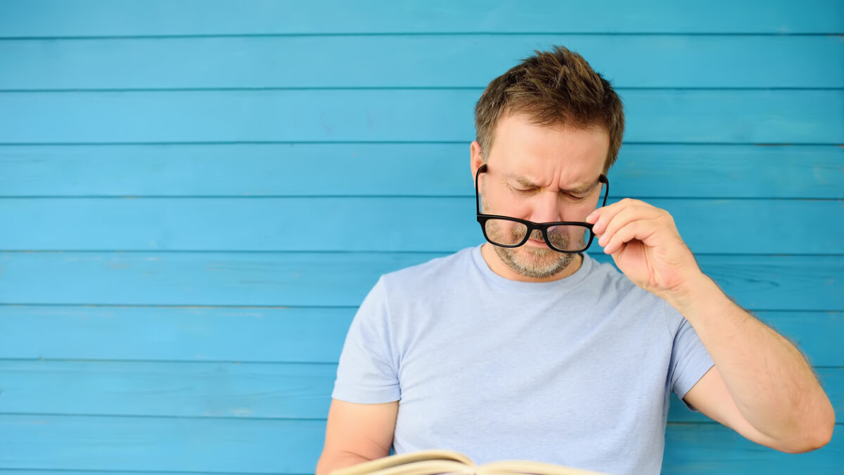 Portrait of mature man with big black eye glasses trying to read book but having difficulties seeing text because of vision problems. Problems disorder vision.