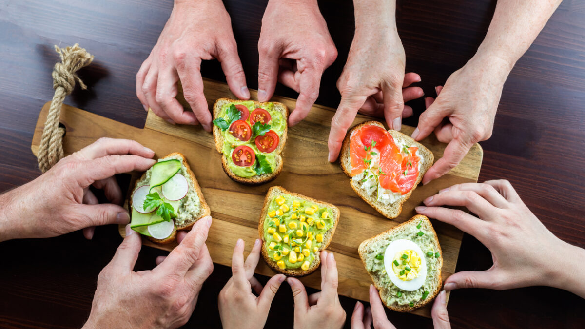 Many hands of different ages of people reach out and hold various healthy and delicious sandwiches. Slices of bread are spread with cottage cheese and avocado sauce, various vegetables on top.