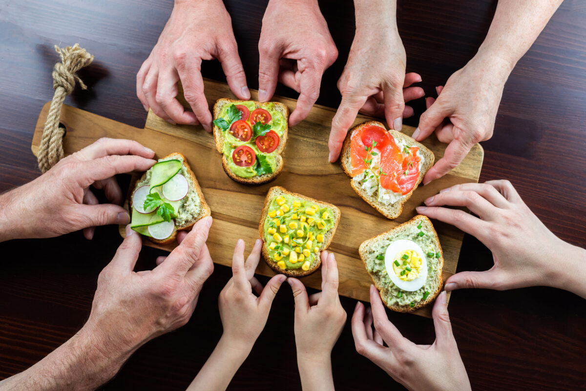 Many hands of different ages of people reach out and hold various healthy and delicious sandwiches. Slices of bread are spread with cottage cheese and avocado sauce, various vegetables on top.
