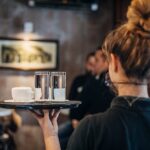 Group of people, female waitress working at cafe, wearing a protective mask. She is holding a trey with coffee cups