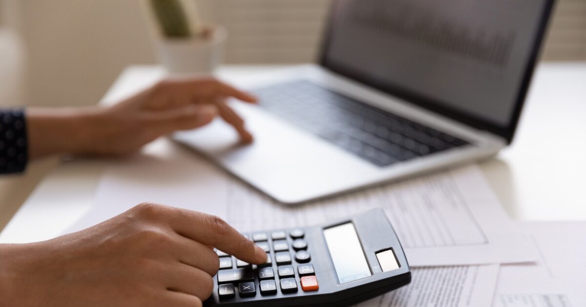 Close up of young female electronic bank client using calculator in paperwork with accounts bills before providing payment online by laptop. Woman hands at work table with papers accounting sum to pay