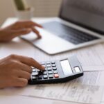 Close up of young female electronic bank client using calculator in paperwork with accounts bills before providing payment online by laptop. Woman hands at work table with papers accounting sum to pay