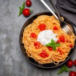 Stracciatella with spaghetti and tomatoes in black dish on dark grey background