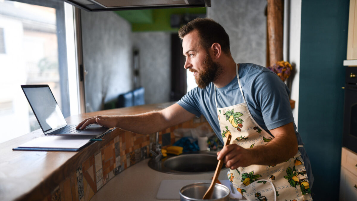 Bearded Male Working On Laptop And Making Dinner At Same Time
