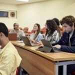 Tiered rows of desks and people in teens, 20s, and 30s smiling, talking, and using their portable devices. Property release attached