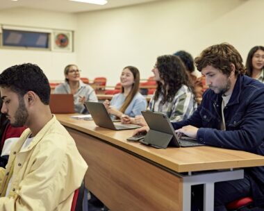 Tiered rows of desks and people in teens, 20s, and 30s smiling, talking, and using their portable devices. Property release attached