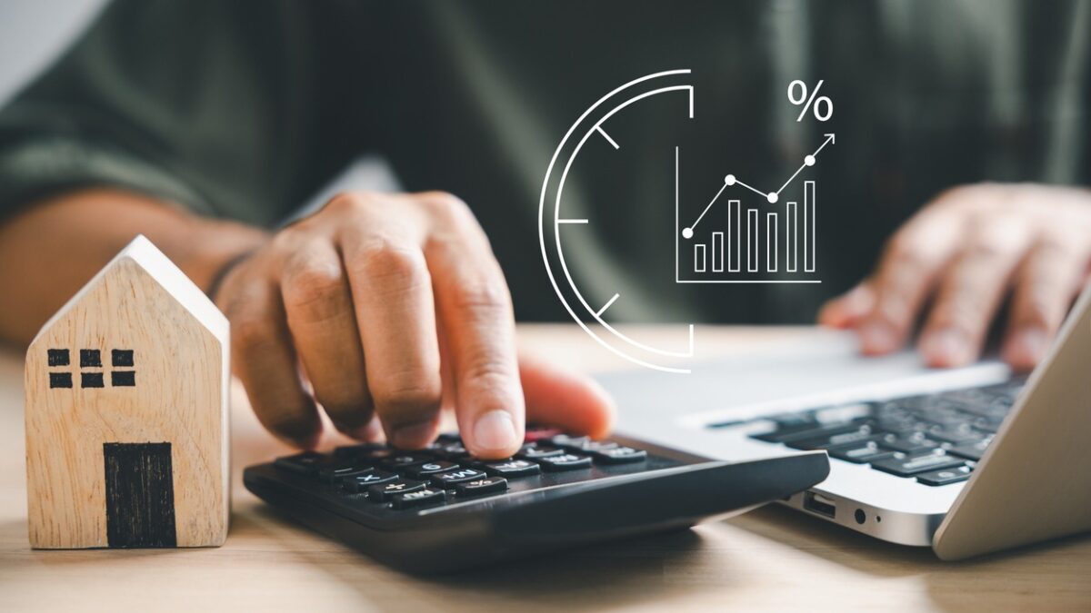 Hand using calculators, planning home refinance. Wooden house model, buy or rent note, calculators on desk. Saving for property purchase, mortgage payment. Tax, credit analysis for financial benefit.