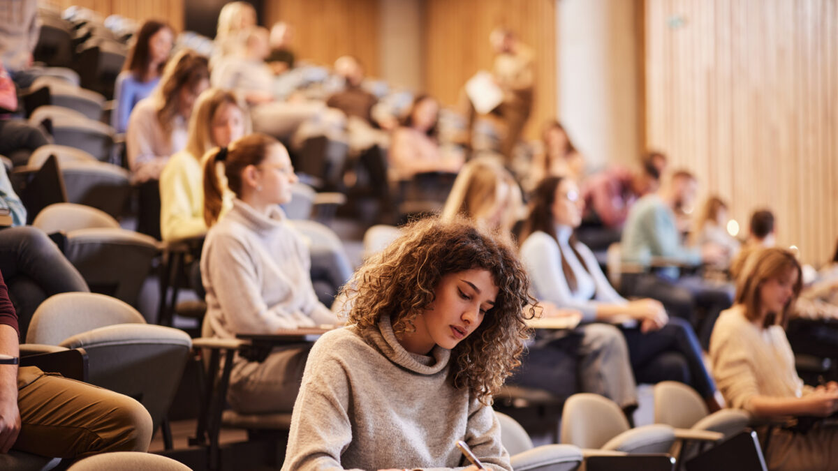 Female college student writing an exam during a class at lecture hall. Her classmate are in the background.
