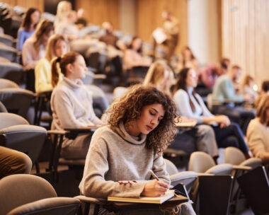 Female college student writing an exam during a class at lecture hall. Her classmate are in the background.