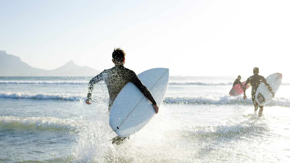 Three teenage boys wearing their wetsuits carrying their surfboards run into the sea on a sunny Summers day
