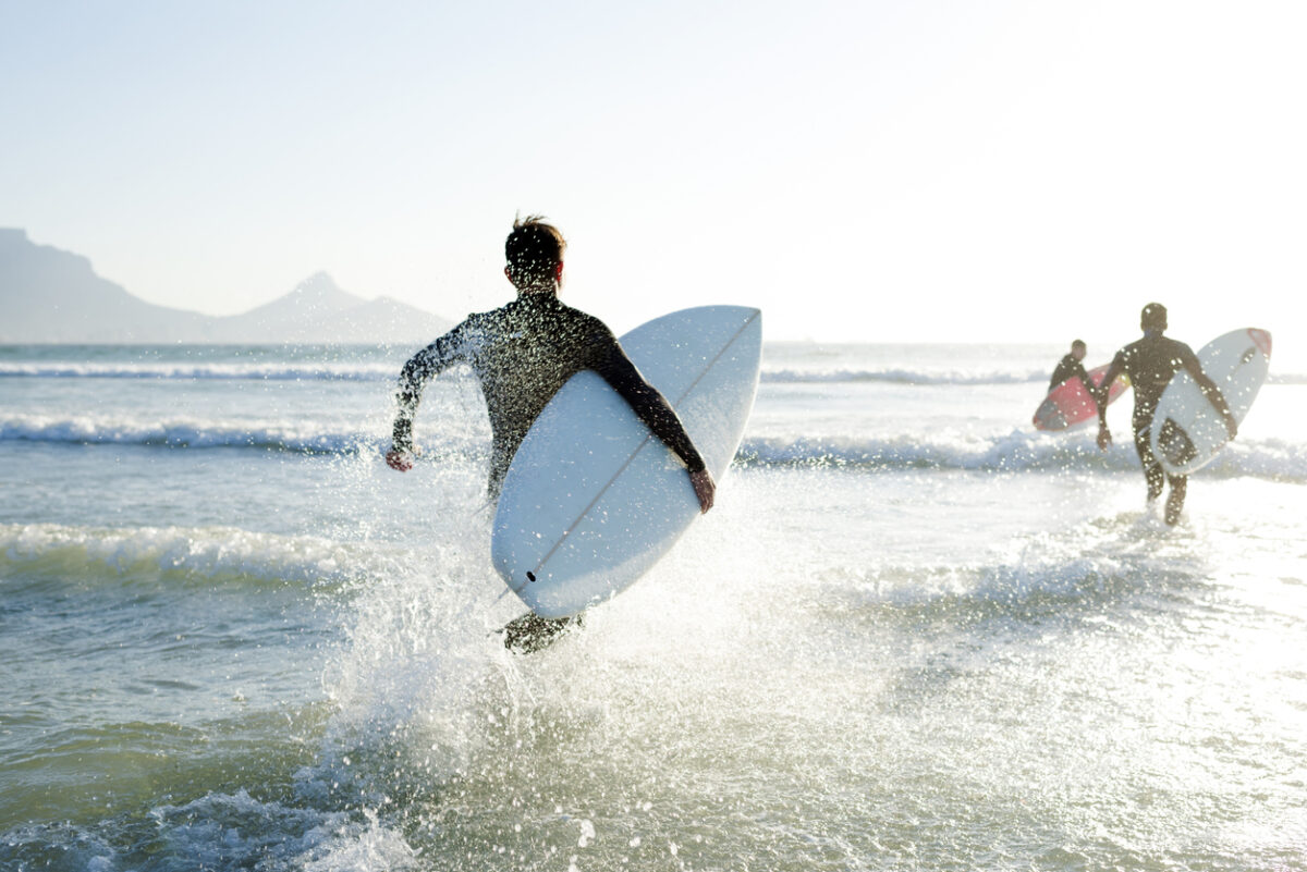 Three teenage boys wearing their wetsuits carrying their surfboards run into the sea on a sunny Summers day