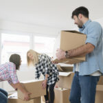 Young man carrying a heavy cardboard box while his female friends carrying one box together.