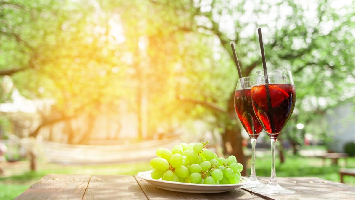 Two glasses of red wine and green grapes stand on wooden table against background of trees and garden in summer, alcoholic cocktails with wine and snack for aperitif outdoors