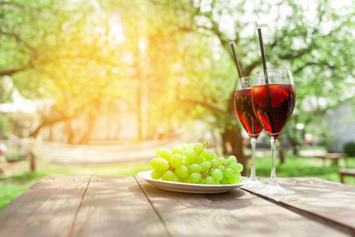 Two glasses of red wine and green grapes stand on wooden table against background of trees and garden in summer, alcoholic cocktails with wine and snack for aperitif outdoors