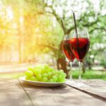 Two glasses of red wine and green grapes stand on wooden table against background of trees and garden in summer, alcoholic cocktails with wine and snack for aperitif outdoors