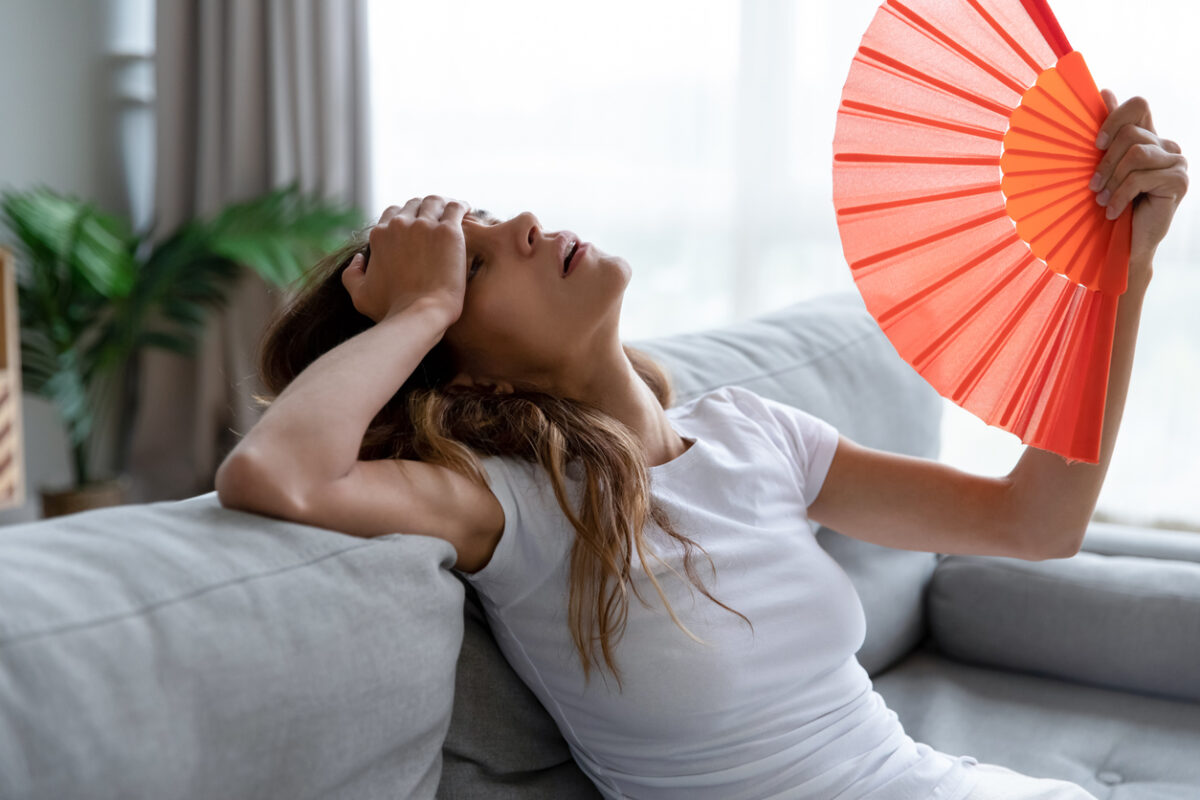 Side view exhausted young biracial girl using paper waver, suffering from hot summer weather or high temperature at home. Unhappy overheated millennial mixed race woman feeling uncomfortable indoors