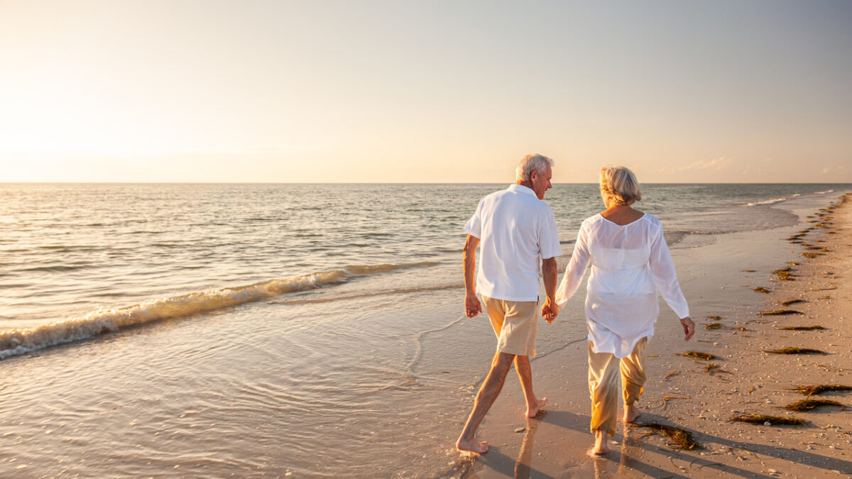 Happy senior man and woman old retired couple walking and holding hands on a beach at sunset