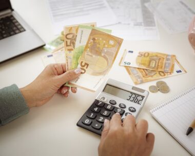Close-up of woman's hands using calculator for planning home finances. She is holding Euro banknotes.