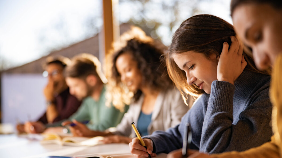 Smiling female student writing an exam while attending a class at college classroom.