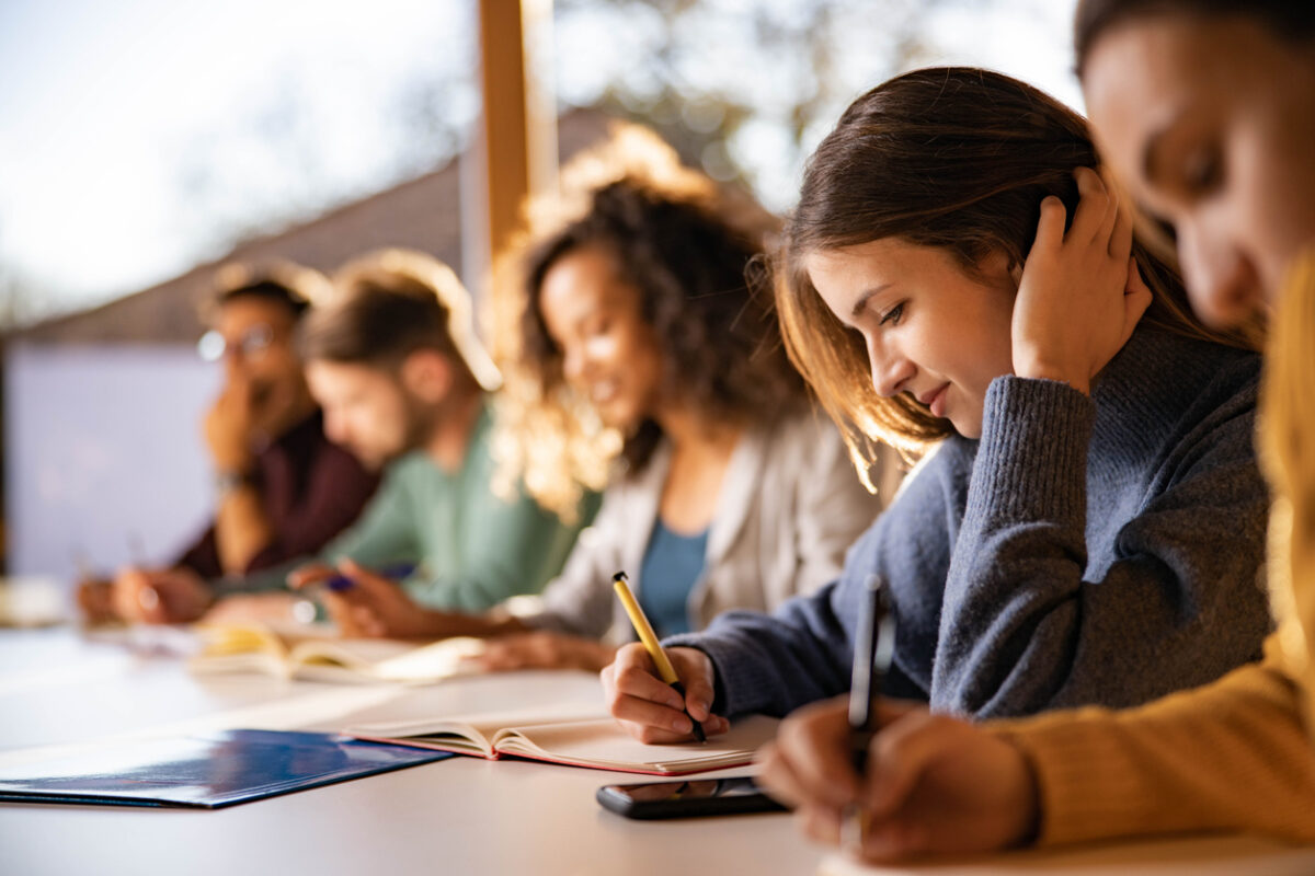 Smiling female student writing an exam while attending a class at college classroom.
