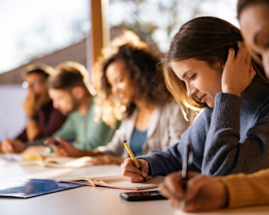Smiling female student writing an exam while attending a class at college classroom.