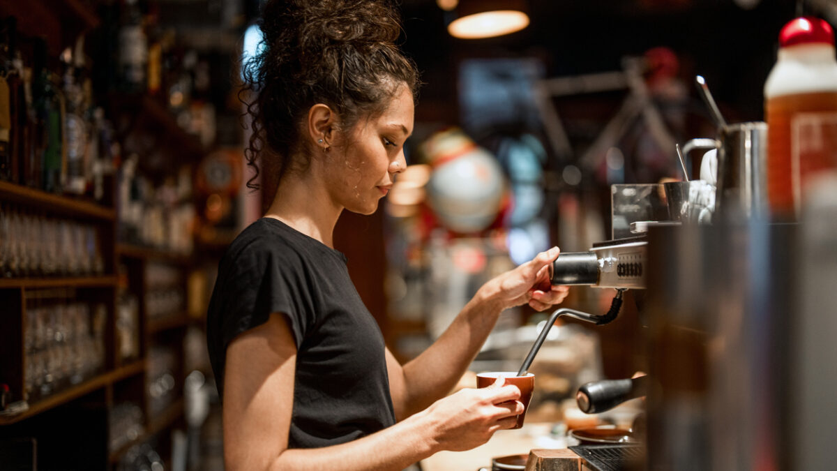 Aboriginal waitress in black attire operates professional espresso machine, demonstrating skilled coffee preparation techniques in a warmly lit cafe environment with traditional bar setup.