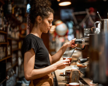 Aboriginal waitress in black attire operates professional espresso machine, demonstrating skilled coffee preparation techniques in a warmly lit cafe environment with traditional bar setup.
