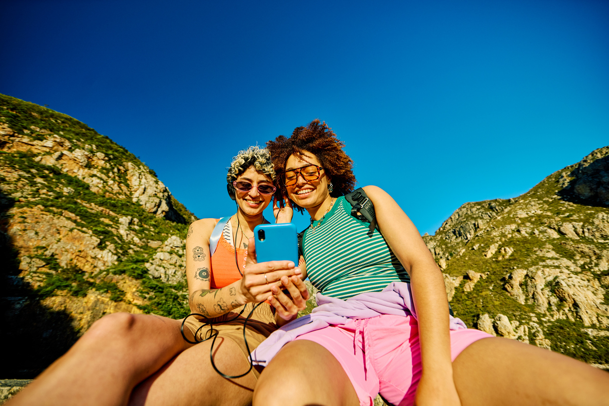 Two young female hikers are taking a break, sitting and listening to music on a smartphone during a mountain trek