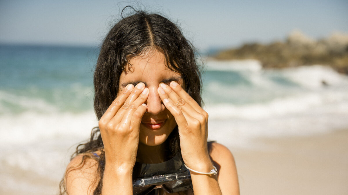 Young woman with diving mask around her neck is standing on a sunny beach, covering her eyes with her hands, likely after a snorkeling session. travel concept