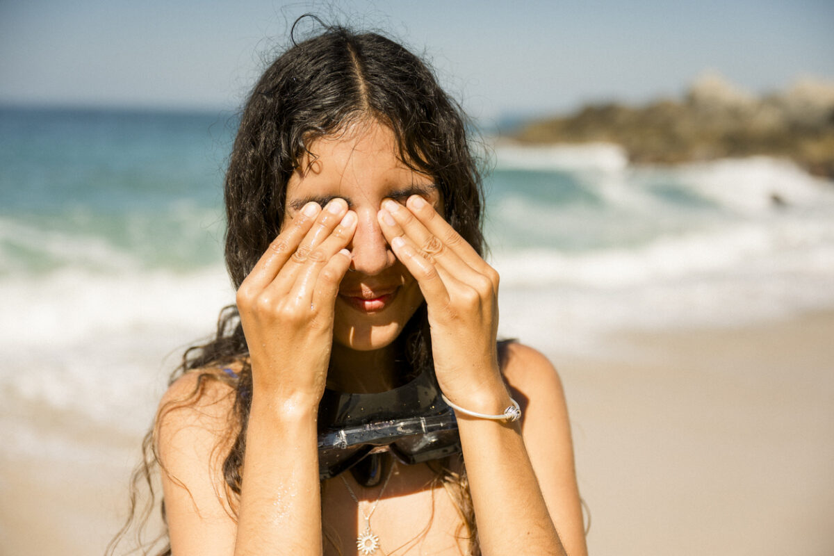 Young woman with diving mask around her neck is standing on a sunny beach, covering her eyes with her hands, likely after a snorkeling session. travel concept