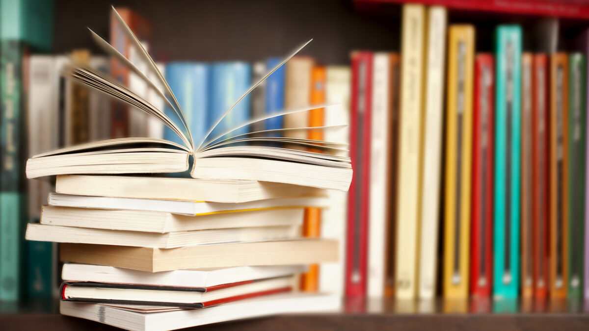 Stack of books on a wooden library shelf, one of them open on top, multicolored book spines background