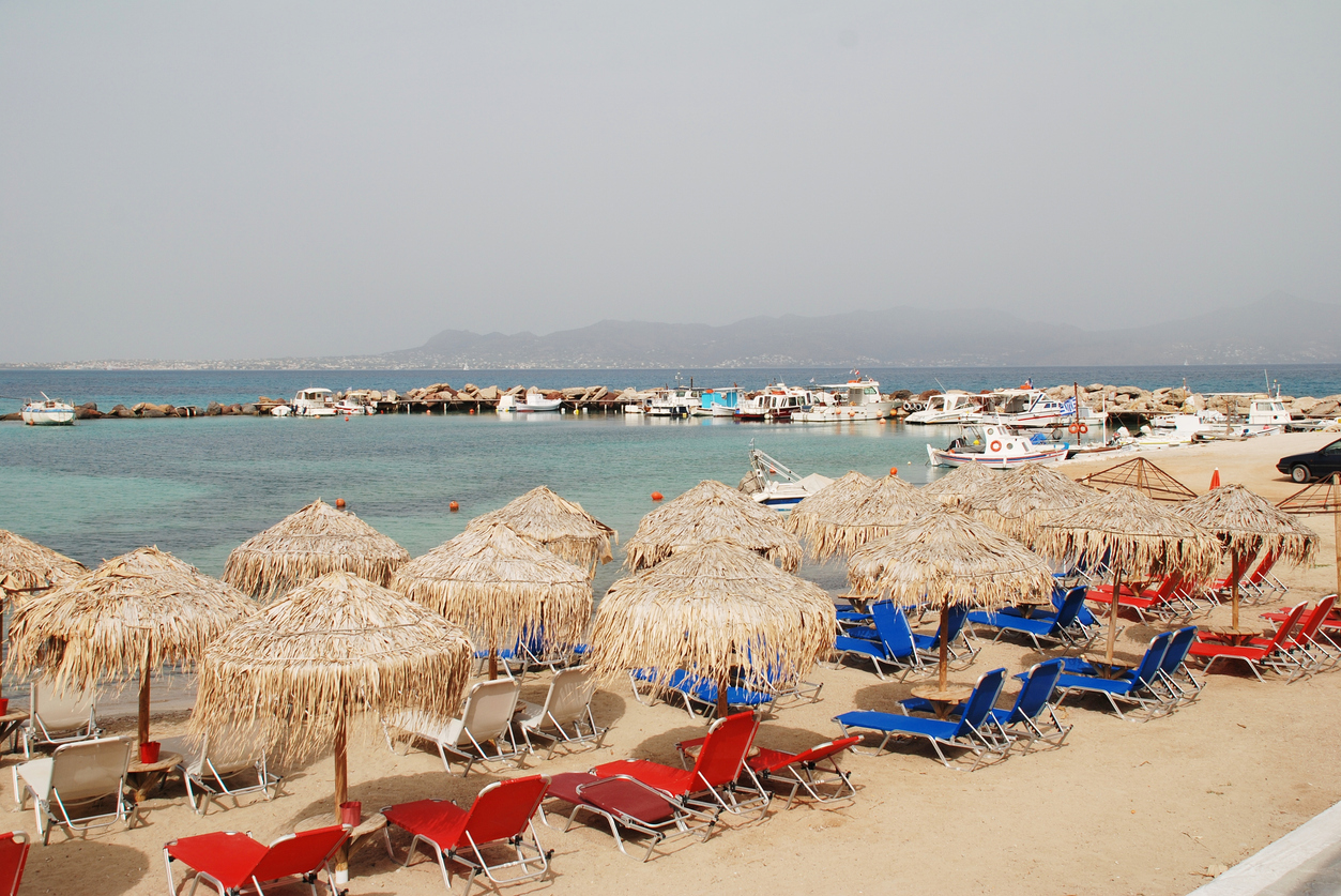 Agistri, Greece - May 12, 2016: The sandy beach at Skala on the Greek island of Agistri. The 5.5km long Saronic island is under an hour from the Athens port of Piraeus. Aegina island is in the background.