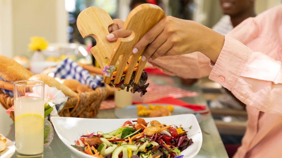 Side view mid section of a biracial woman using salad tongs to serve herself from a bowl of salad, sitting at a table during a family meal