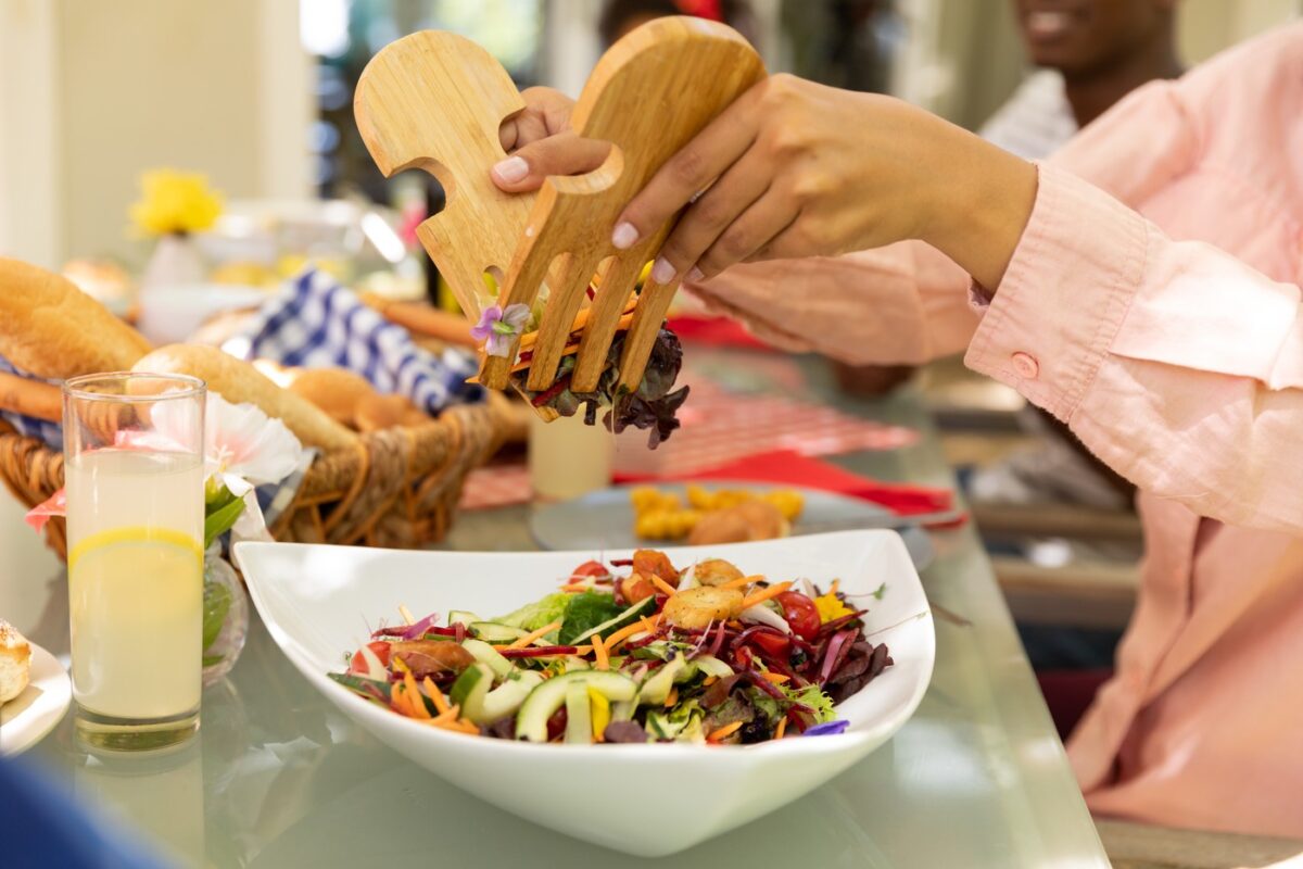 Side view mid section of a biracial woman using salad tongs to serve herself from a bowl of salad, sitting at a table during a family meal