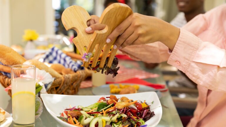 Side view mid section of a biracial woman using salad tongs to serve herself from a bowl of salad, sitting at a table during a family meal