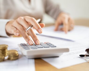 Woman using a calculator doing a balance of monthly expenses. Close up of businesswoman working with financial data and using calculator with coins on desk at office. Deatil of hands calculating home finances and annual taxes