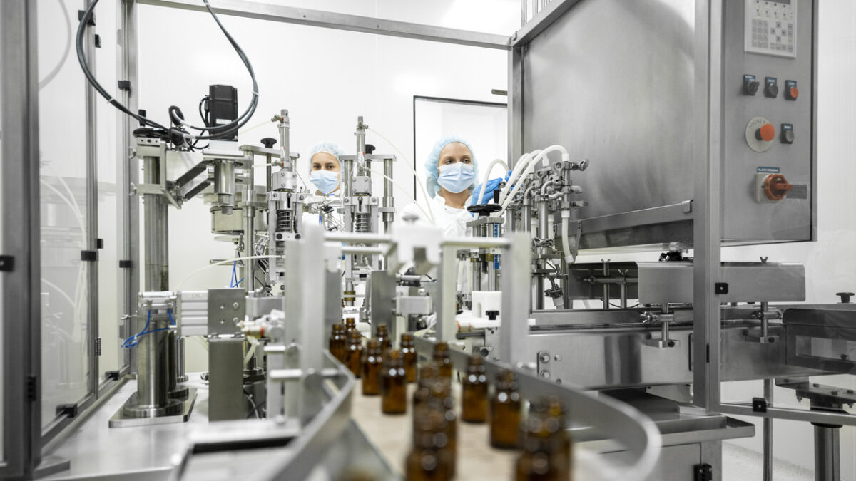 Small brown bottles seen perfectly arranged in a laboratory machine during manufacturing in a pharmaceutical factory while two female employees in a protective clothing are controlling the process