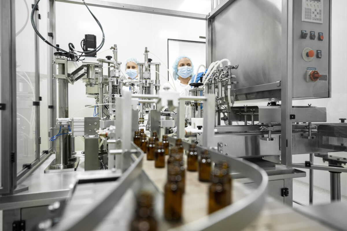 Small brown bottles seen perfectly arranged in a laboratory machine during manufacturing in a pharmaceutical factory while two female employees in a protective clothing are controlling the process