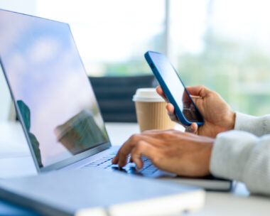 Close up of a Businessman working on a laptop computer and holding and looking at a mobile phone in the office. There are notepads on the desk. He is casually dressed