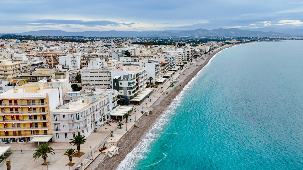 An aerial view of Loutraki, a charming coastal town in Greece known for its scenic waterfront promenade and vibrant urban landscape. The image highlights the turquoise waters of the Corinthian Gulf, lined with modern buildings, green parks, and palm trees, creating a perfect blend of natural beauty and urban charm. Loutraki is renowned for its thermal spas, cultural heritage, and inviting beaches, making it a popular destination for tourists seeking relaxation and leisure. The town's unique location provides breathtaking views of the sea and surrounding mountains, ideal for travel and lifestyle themes.