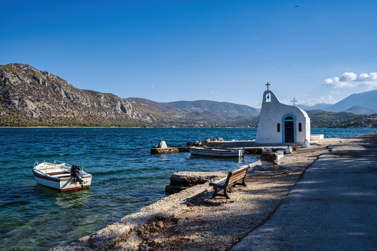 A small white chapel with a bell tower stands on the shore of Limni Vouliagmenis in Loutraki, Greece, surrounded by boats and a scenic mountain backdrop.
