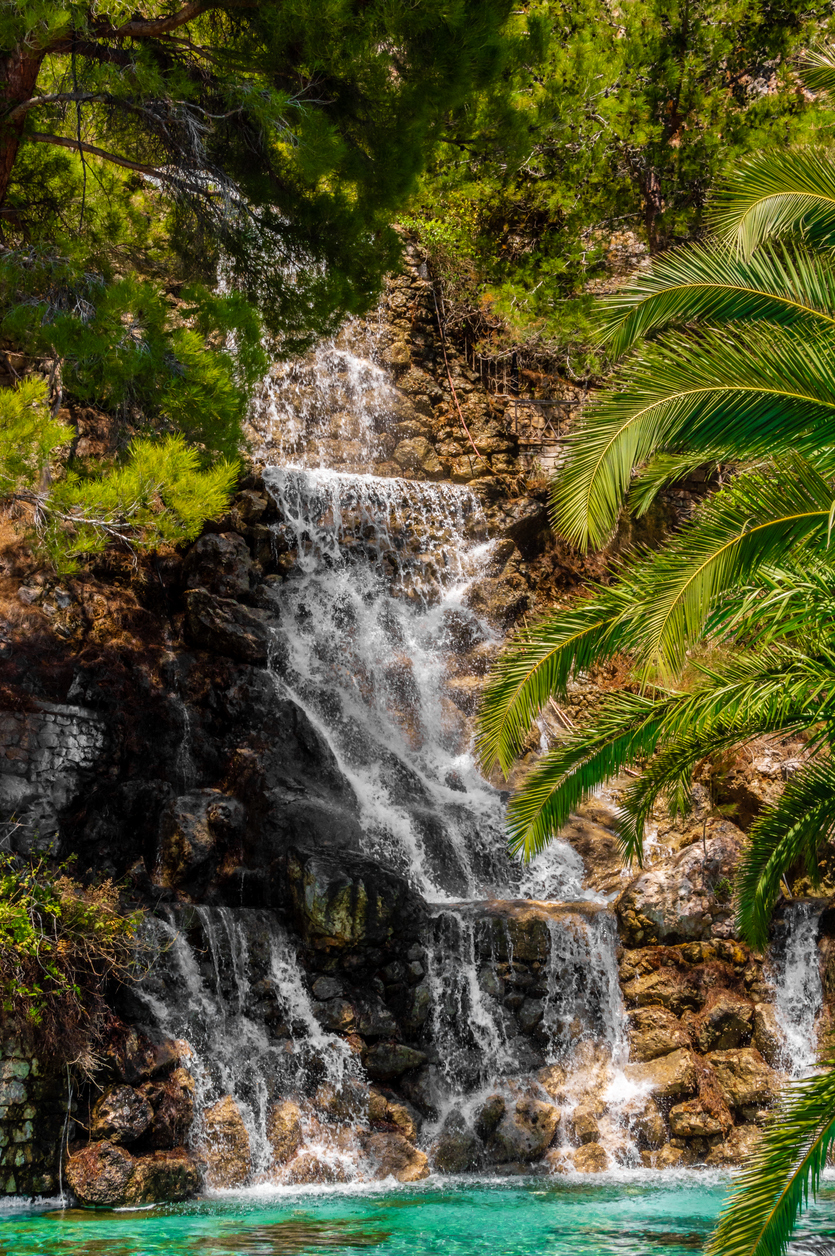 A vibrant waterfall cascades over rocky stones surrounded by lush greenery and palm trees in Loutraki, Corinth, Greece. A peaceful summer scene in bright sunlight.